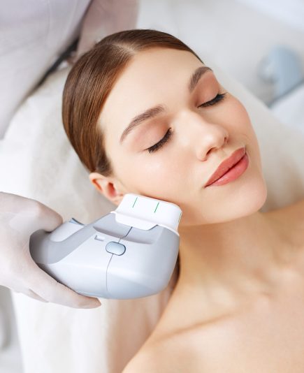 From above of relaxed young female client getting SMAS ultrasound face lifting massage with professional equipment in beauty center