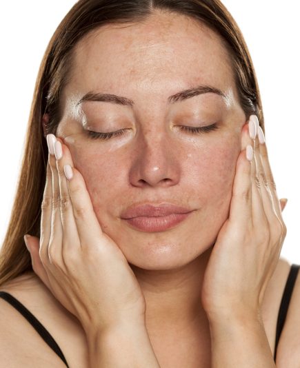 beautiful middle-aged woman applying moisturizer on her face on white background