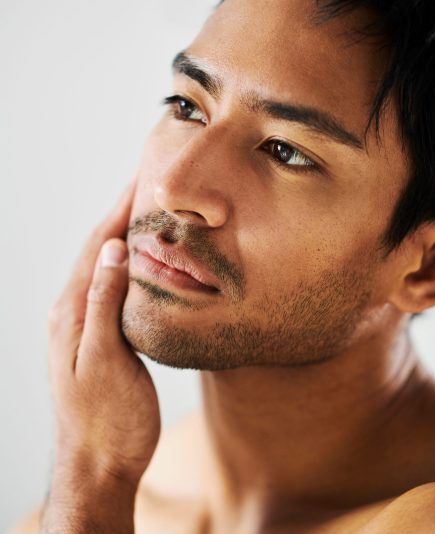 Close up of attractive young Indian male with stubble touching face and looking at skin. Metrosexual man examining complexion. The results of a good facial skincare or shaving regime