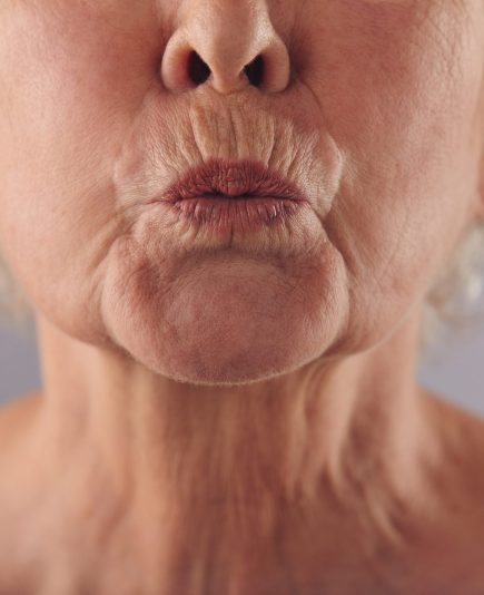 Close-up portrait of mature woman puckering lips against grey background. Senior woman grimacing. Focus on lips.