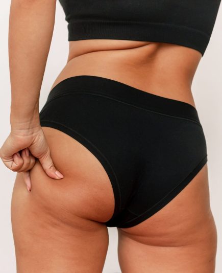 Cropped shot of a young tanned woman in black panties grabbing skin on her buttock with excess fat isolated on a white background. Buttocks and thighs with cellulite. Excess weight, overweight